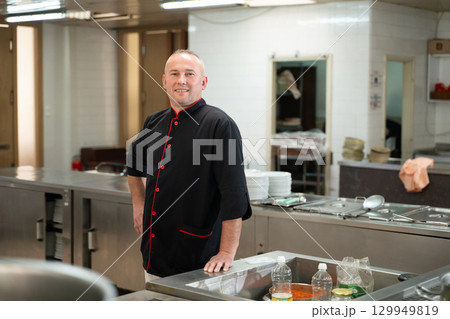 Portrait of a male chef in a black uniform with red details standing in a professional restaurant kitchen. Image highlights hospitality industry, confidence, and culinary expertise Portrait of a male chef in a black uniform with red details standing in a professional restaurant kitchen. Image highlights hospitality industry, confidence, and culinary expertise 129949819