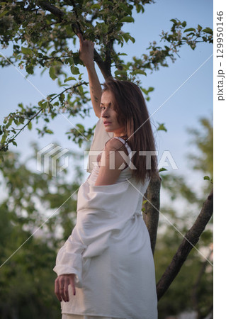 A woman in a white outfit stretches her arms upwards, leaning against a tree. This image showcases the peaceful and harmonious interaction with the natural environment. 129950146