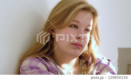 young woman with a melancholic expression is bathed in natural light while leaning against a wall. The scene conveys themes of emotional depth and personal reflection. 129950150