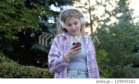 A teenage girl checks her smartphone while wearing headphones in a peaceful park setting. The image captures the essence of modern connectivity and outdoor tranquility. A teenage girl checks her smartphone while wearing headphones in a peaceful park setting. The image captures the essence of modern connectivity and outdoor tranquility. 129950162
