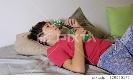 The boy gazes thoughtfully while lying down, embracing a moment of calm with his toy guitar. The boy gazes thoughtfully while lying down, embracing a moment of calm with his toy guitar. 129950173
