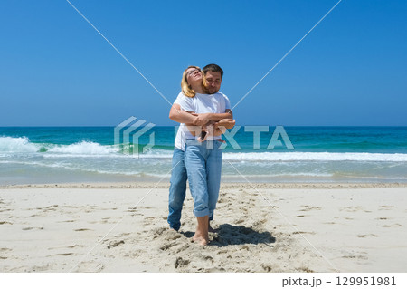 Couple standing barefoot on sandy beach with man hugging woman from behind in joyful moment. 129951981