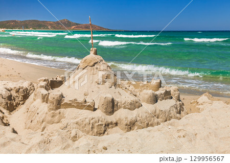Detailed sandcastle on sunny Sardinian beach with turquoise waves and coastal hills in the background 129956567