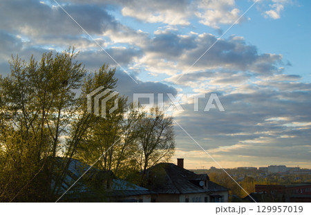 Roof of the old house behind trees against cloudy sky with clouds and sunset 129957019