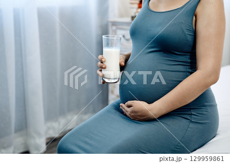 Close-up of pregnant woman in blue dress sitting on bed holding a glass of milk and touching belly with copy space 129959861