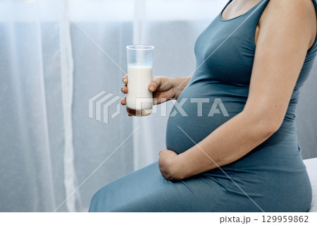 Close-up of pregnant woman in blue dress holding glass of milk and touching belly indoors with copy space Close-up of pregnant woman in blue dress holding glass of milk and touching belly indoors with copy space 129959862