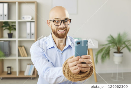 Crazy man holding smartphone with hands tied by rope, having addiction to phone and social media. 129962309