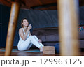 smiling woman sitting on yoga mat at home drinking water after exercising 129963125