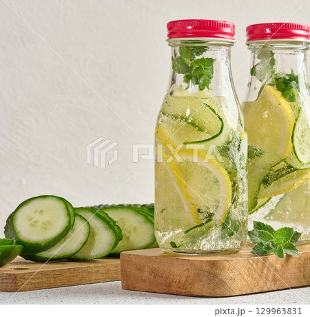 Glass bottles with red lids, filled with detox water containing cucumber and lemon slices and mint leaves, white table Glass bottles with red lids, filled with detox water containing cucumber and lemon slices and mint leaves, white table 129963831