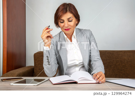 Professional woman reviewing notes while seated at a desk with digital devices 129964274