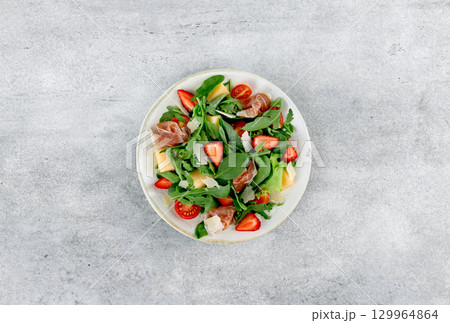 Fresh Italian salad with arugula, strawberries, melon,prosciutto,cheese on gray background. Top view 129964864