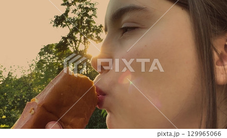 Big bite of a burger in the park, Girl wears a striped t-shirt, Sunlight glimmers through trees, She enjoys every chew, Lush greenery surrounds, Outdoor lunch feels amazing, Smile lights up her face 129965066