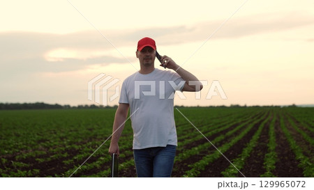 Agriculture, Rural Farmer Communicating, Man wearing a red hat, talking on mobile in a green field, casual day on the farm, relaxed stance, young farmer, fields stretching far, checking crop Agriculture, Rural Farmer Communicating, Man wearing a red hat, talking on mobile in a green field, casual day on the farm, relaxed stance, young farmer, fields stretching far, checking crop 129965072