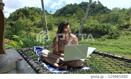 Young woman enjoys the tranquility of nature while working on her laptop in a hammock, surrounded by lush greenery and rice fields  129965880