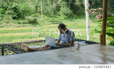 A young man relaxes in a hammock on a bamboo terrace, typing on his laptop, surrounded by lush greenery, tranquility of remote work in tropical setting  129965881