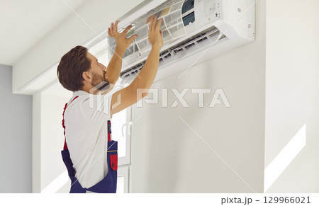 Male professional technician cleans or replaces air conditioner filter hanging on wall inside house. 129966021