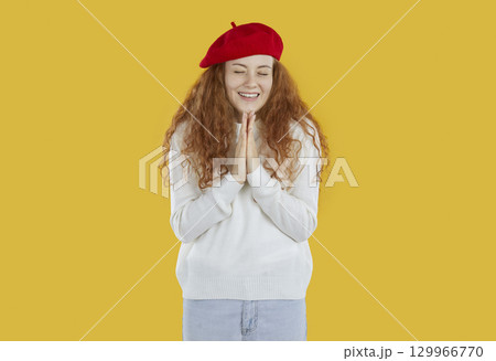 Smiling young redhead girl in white sweater folded hands in prayer on yellow background. 129966770