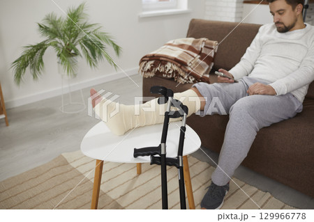 Man with a broken leg sitting on the sofa, with his foot resting on a stool, with crutches beside Man with a broken leg sitting on the sofa, with his foot resting on a stool, with crutches beside 129966778