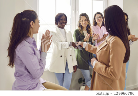 Two businesswomen shaking hands while colleagues applauding them in office 129966796