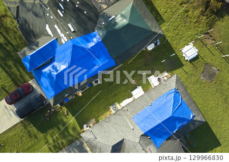 Top view of leaking house roof covered with protective tarp sheets against rain water leaks until replacement of asphalt shingles. Damage of building rooftop as aftermath of hurricane Ian in Florida Top view of leaking house roof covered with protective tarp sheets against rain water leaks until replacement of asphalt shingles. Damage of building rooftop as aftermath of hurricane Ian in Florida 129966830