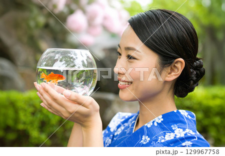 青地に白い花柄の浴衣を着た女性が、庭園で金魚鉢を見つめ微笑む夏の情景 129967758