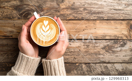 木製のテーブルの上でラテアートコーヒーを持つ女性の手元 129968313