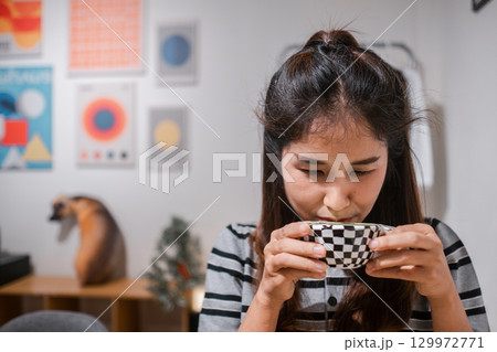 A woman sipping a hot drink in a stylish room with geometric art posters, creating a cozy and modern ambiance. A woman sipping a hot drink in a stylish room with geometric art posters, creating a cozy and modern ambiance. 129972771