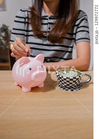 A woman places a coin in a piggy bank while enjoying a matcha latte, symbolizing savings and relaxation. A woman places a coin in a piggy bank while enjoying a matcha latte, symbolizing savings and relaxation. 129972772