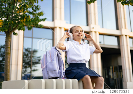 Caucasian schoolgirl in a schoolyard wearing headphones and listening to music, with a backpack. Caucasian schoolgirl in a schoolyard wearing headphones and listening to music, with a backpack. 129973525