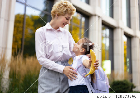 A mom and her daughter share a tender moment in a schoolyard, with the little girl holding a banana. A mom and her daughter share a tender moment in a schoolyard, with the little girl holding a banana. 129973566