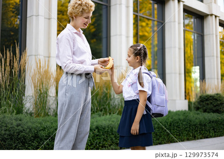 A mother gives a banana to her daughter in a schoolyard. The daughter wears a backpack and school uniform. 129973574