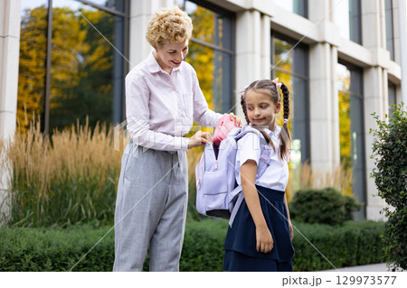 A mother helps her daughter pack her backpack in front of a school building. The little girl is wearing her school uniform. A mother helps her daughter pack her backpack in front of a school building. The little girl is wearing her school uniform. 129973577