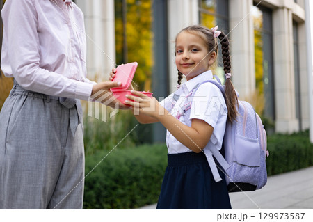 A mom hands her daughter a lunchbox at the schoolyard, preparing her for the school day. 129973587