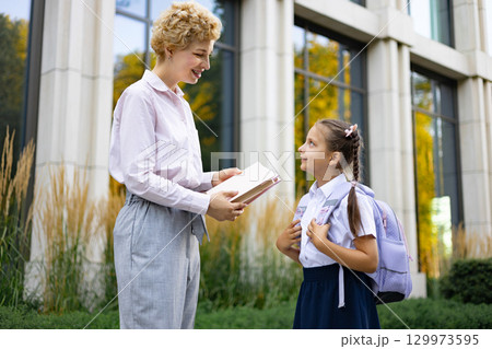 A mother smiles as she speaks to her daughter in a schoolyard, holding a book. A mother smiles as she speaks to her daughter in a schoolyard, holding a book. 129973595