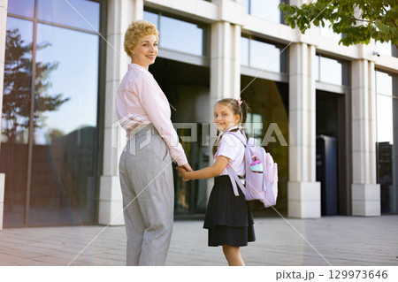 A mother and her elementary school-aged daughter holding hands in front of a school building. 129973646