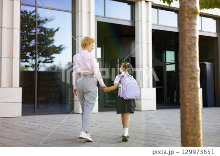 A mother and daughter are walking hand-in-hand toward the school entrance on a sunny day. A mother and daughter are walking hand-in-hand toward the school entrance on a sunny day. 129973651