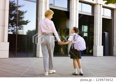 A mother and her daughter holding hands as they walk outside a school building. A mother and her daughter holding hands as they walk outside a school building. 129973652