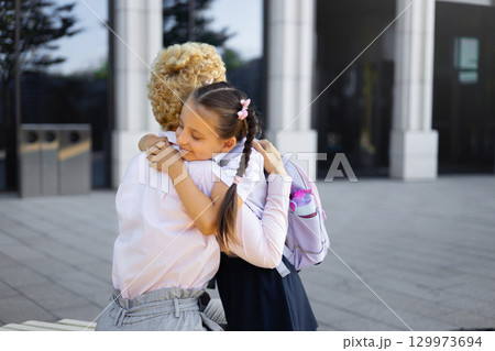 A mom and daughter share a loving hug in front of a school building. The girl wears a school uniform and backpack. 129973694