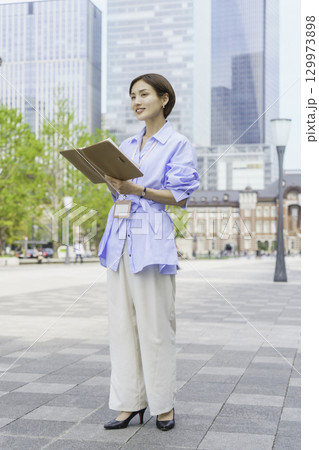 オフィス街で手帳を持って立つ若いビジネスウーマンのポートレート　縦位置 129973898
