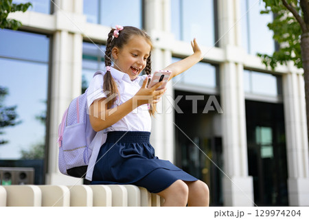 A young school girl with a backpack enthusiastically using her phone on a school campus. 129974204