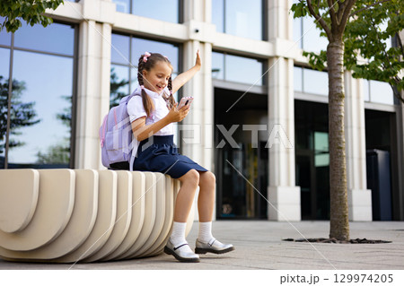 A young schoolgirl in a uniform sits on a bench, smiling while holding a phone, a backpack next to her, on a school yard. A young schoolgirl in a uniform sits on a bench, smiling while holding a phone, a backpack next to her, on a school yard. 129974205