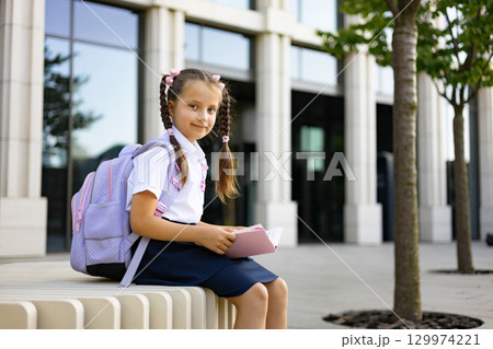 A young girl with pigtails and a backpack sits outside a school, holding a book. A young girl with pigtails and a backpack sits outside a school, holding a book. 129974221
