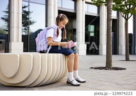 A young schoolgirl with a backpack sits and reads a book on a bench in front of a school building. 129974223