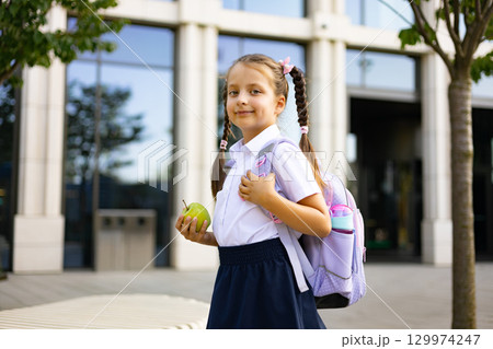 A young schoolgirl with pigtails and a backpack stands on a school yard, holding an apple. 129974247