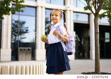 A young girl with pigtails carries a backpack and an apple, smiling at the camera in front of a school building. 129974248