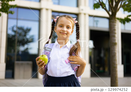A young girl in school uniform, smiling, holding an apple, and wearing a backpack on a school yard. 129974252