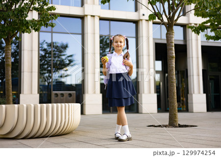 A young schoolgirl stands on the school yard, holding an apple, with a backpack, looking at the camera. 129974254