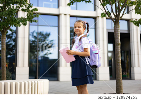 A schoolgirl with a backpack stands in front of the school building, holding a book. 129974255