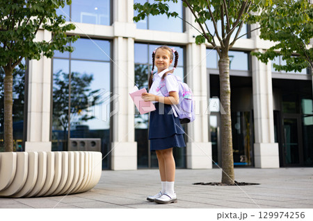 A schoolgirl with pigtails stands outside, holding a book and wearing a backpack, ready for her studies. 129974256