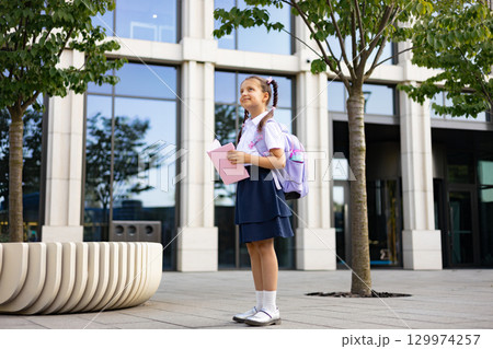 A young girl with braids and a backpack stands on the schoolyard with a book, looking up at the sky. 129974257